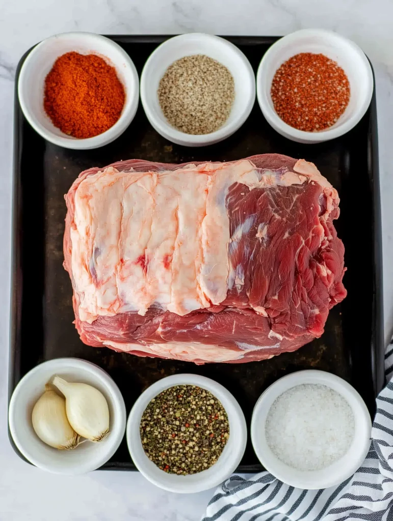 Raw beef roast for Poor Man’s Prime Rib surrounded by small bowls of seasonings, including pepper, salt, garlic, and paprika, neatly arranged on a black tray.